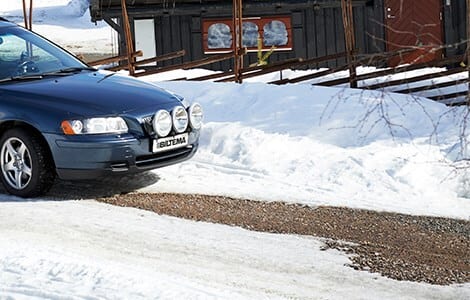 Blue car in snow with gravel path next to a dark building.