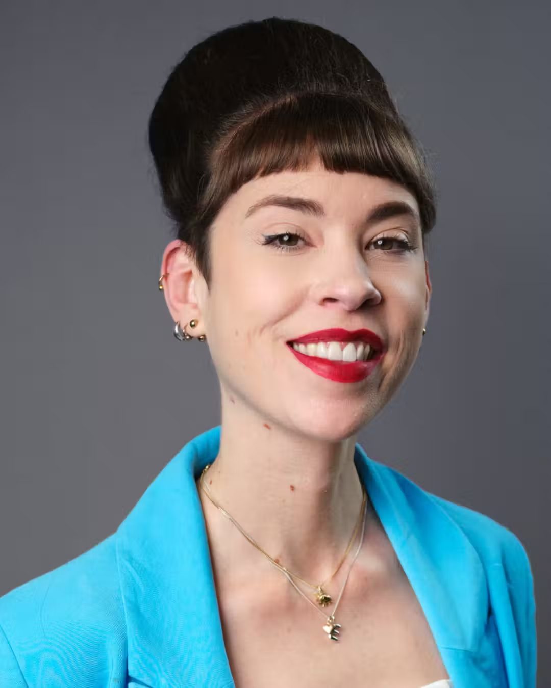 A smiling woman with a beehive hairstyle, bangs, red lipstick, and a blue blazer.