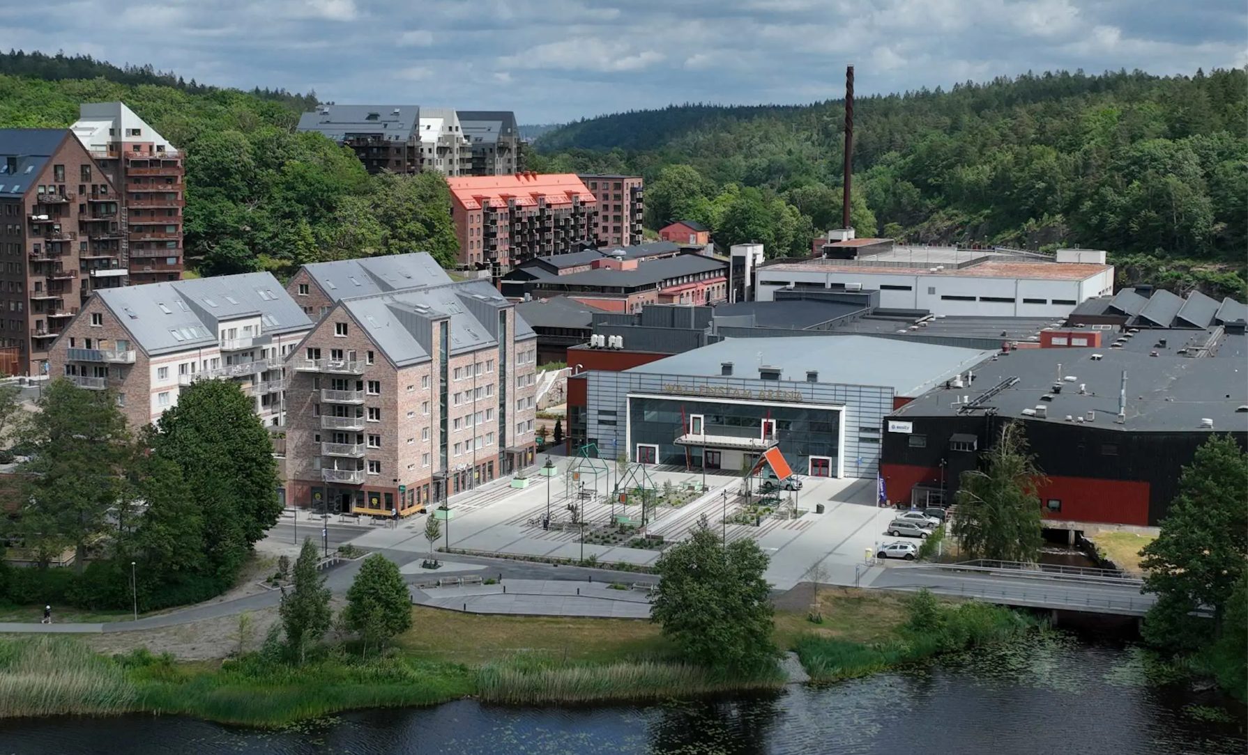 Aerial view of a town with modern buildings, green hills, and a river under a cloudy sky.