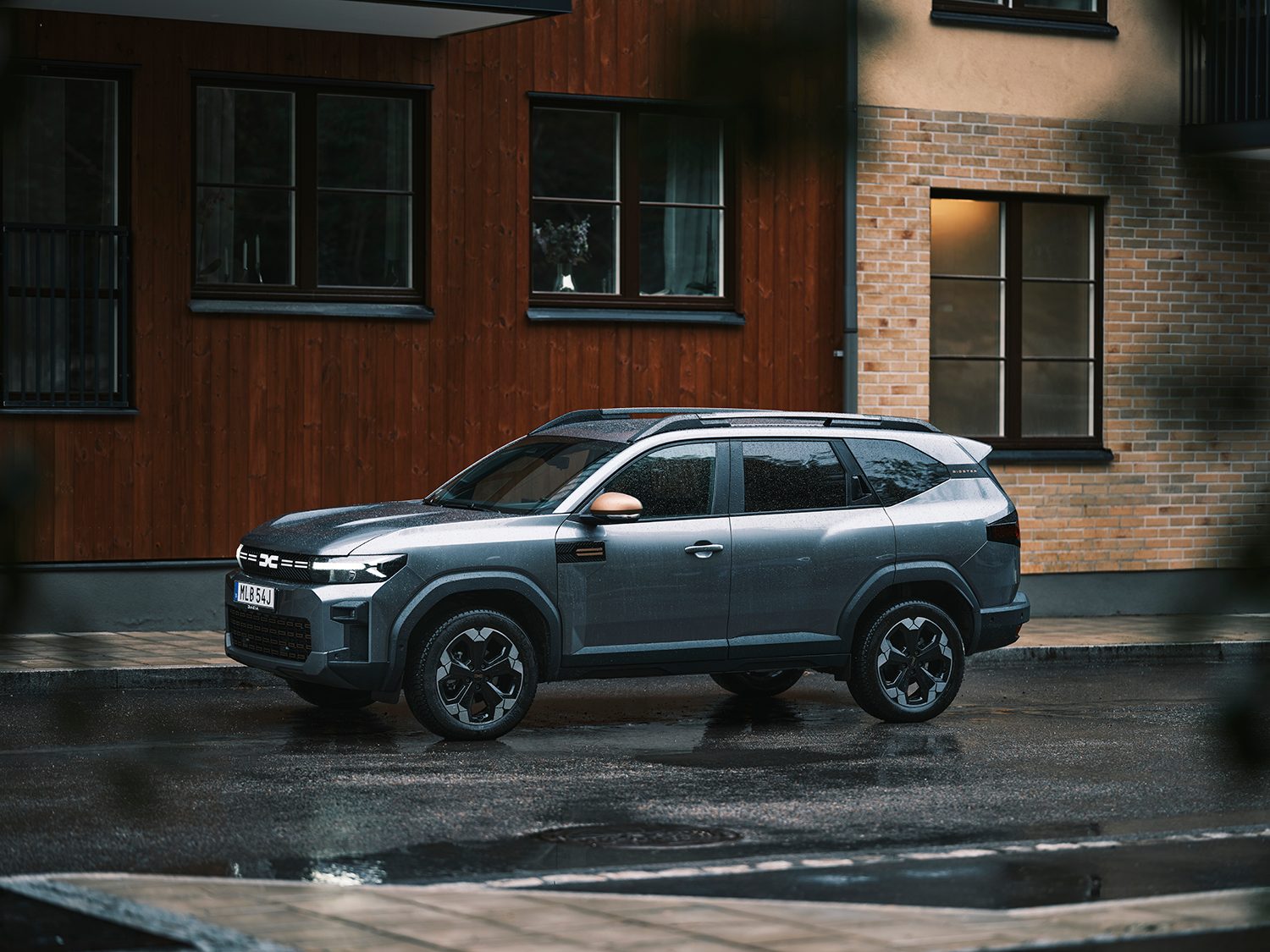 A grey Dacia Duster SUV parked on a wet street in front of residential buildings.