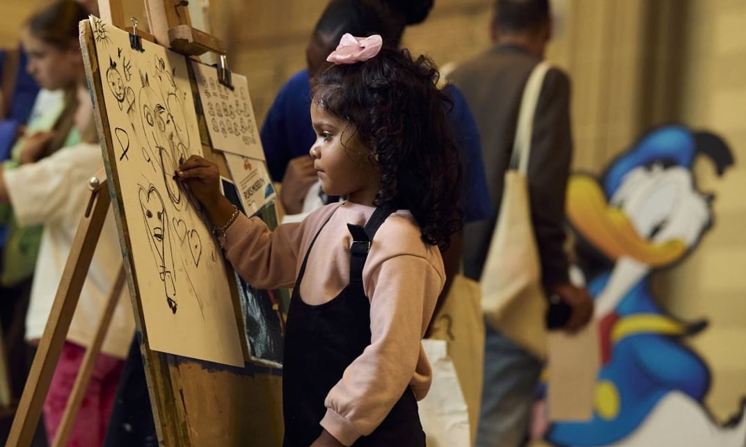 A little girl with curly hair draws on an easel, with a Donald Duck cutout in the background.