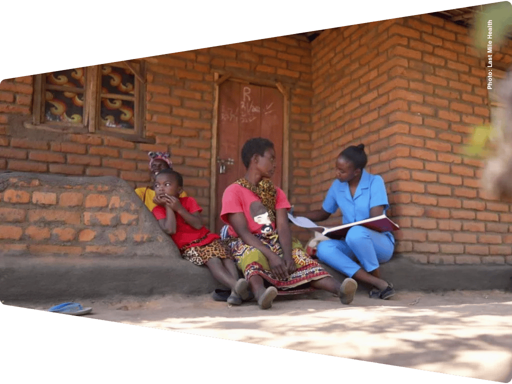 Health worker consults with a mother and child outside a brick home, reviewing paperwork.