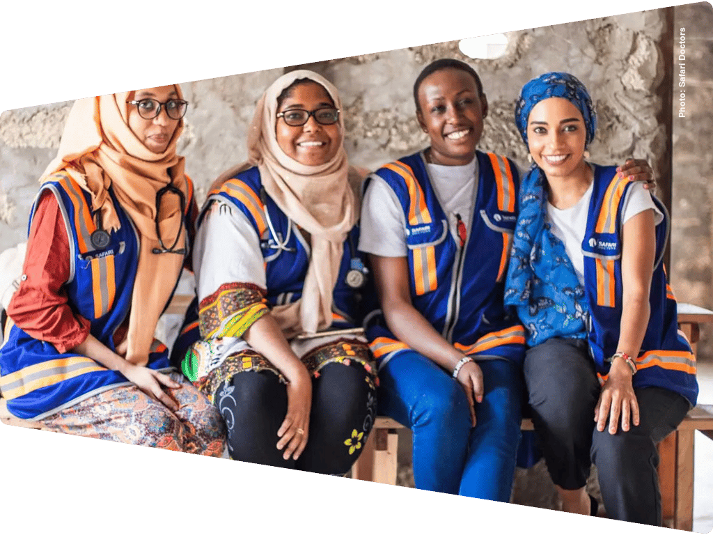 Four smiling women, likely healthcare professionals, wearing blue and orange vests and stethoscopes.