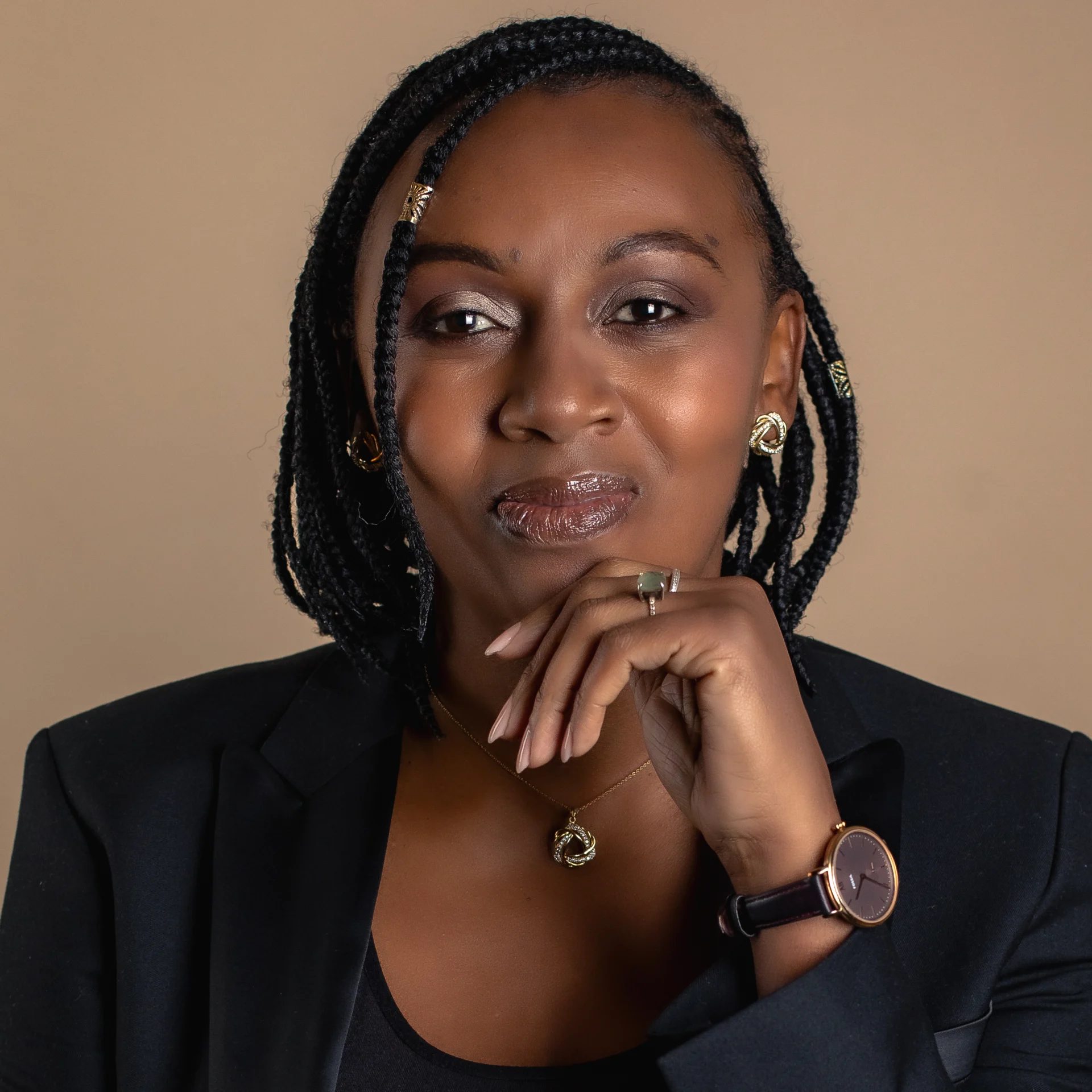 A woman with braided hair and a black blazer, resting her chin on her hand, looking directly at the camera.