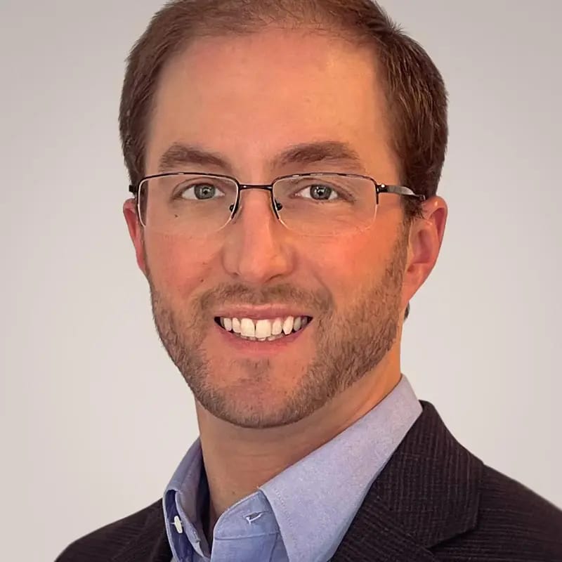 A headshot of a smiling man with glasses, a beard, a blue shirt, and a dark jacket.