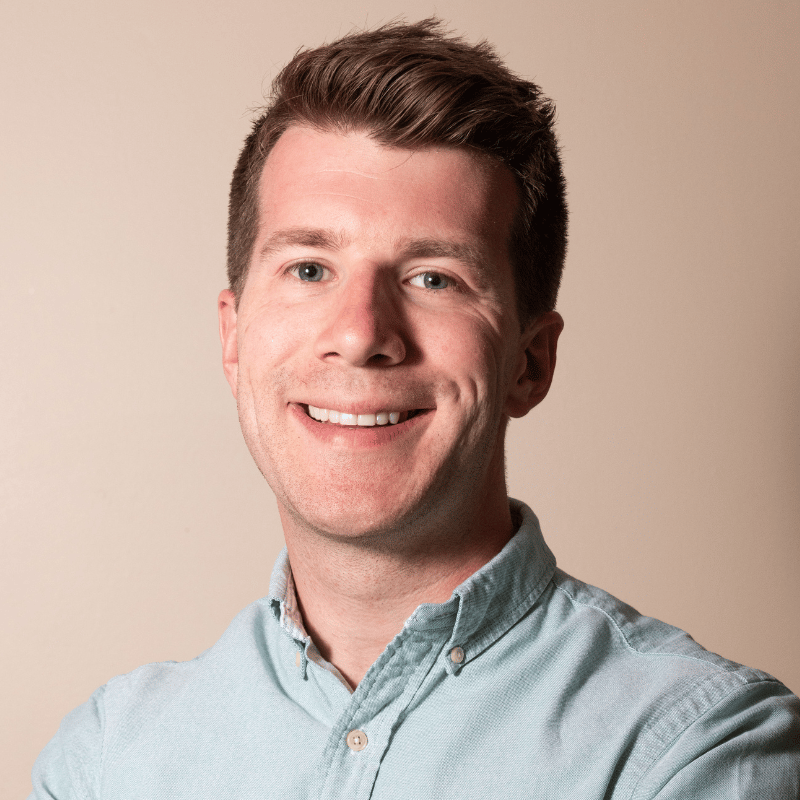 A smiling man with short brown hair and blue eyes wearing a light blue button-up shirt.