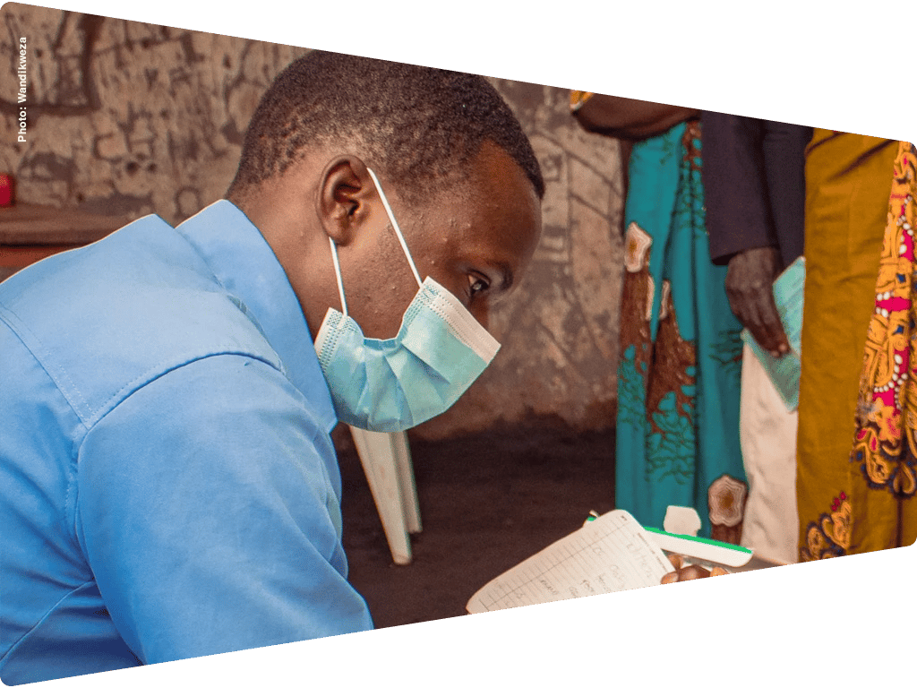 A man in a blue shirt and mask writes in a notebook. Others in traditional attire are in background.