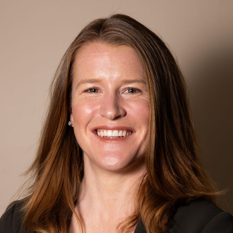 Headshot of a smiling woman with long brown hair, wearing a dark jacket, looking forward.