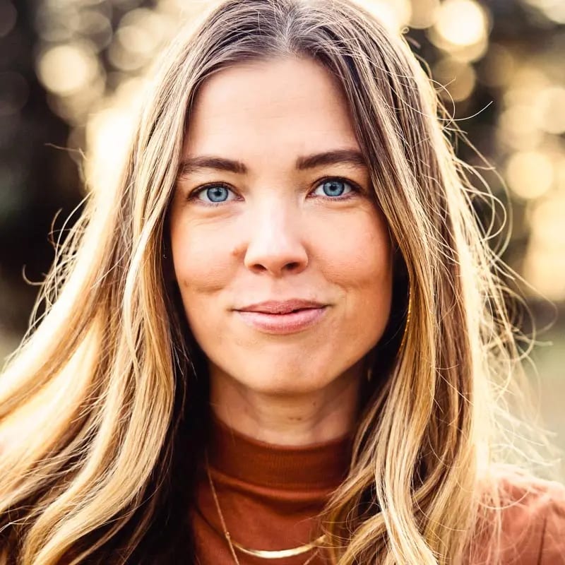Smiling woman with striking blue eyes, long blonde hair, and a brown top, looking at the camera.