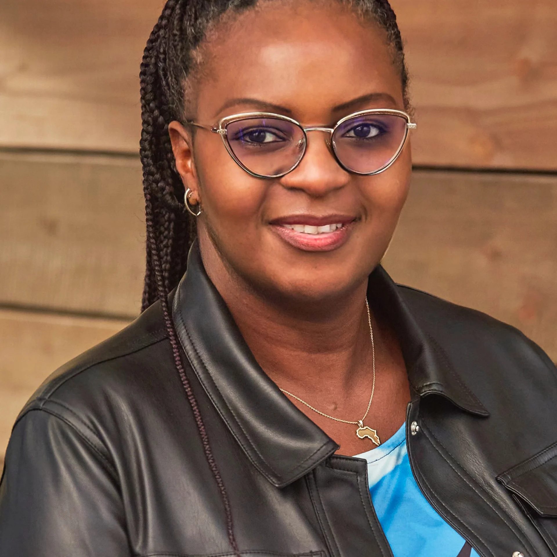 A smiling Black woman with glasses, braided hair, and a black leather jacket, wearing an Africa-shaped necklace.