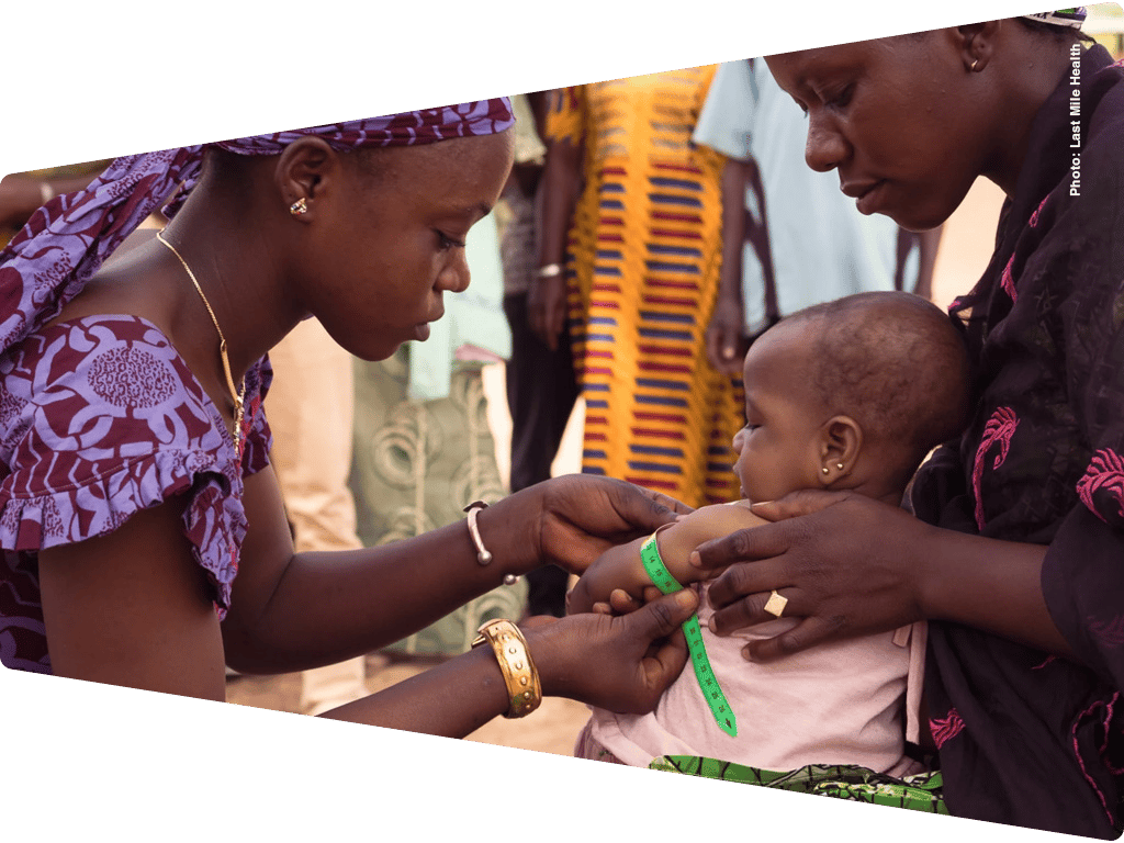 Two women measure a baby's arm with a green tape, assessing health in a community setting.