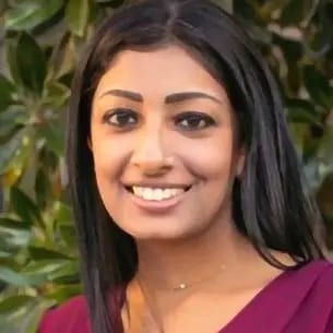 Headshot of a smiling woman with long dark hair, wearing a dark top, against blurred green foliage.