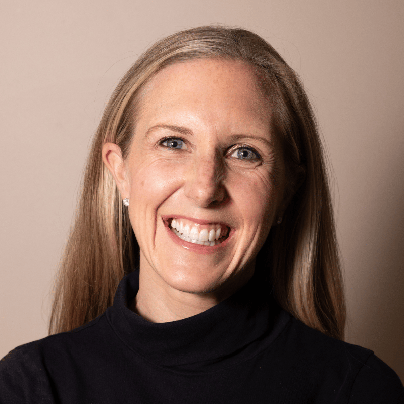 A headshot of a smiling woman with blonde hair and blue eyes, wearing a black turtleneck.