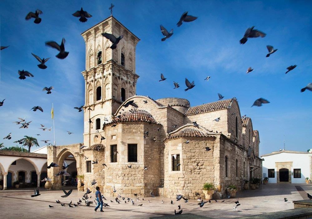 Stone church with a tall bell tower and domes, framed by a blue sky and many flying pigeons.