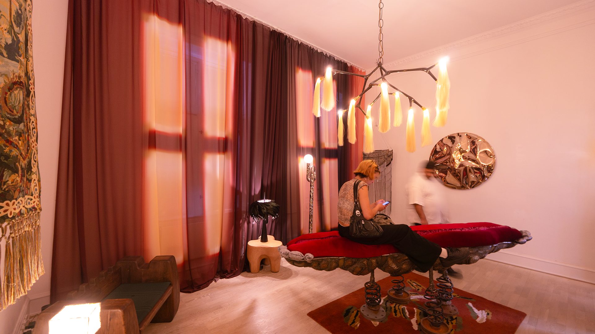 Woman on red bench, tassel chandelier, metallic wall art, sheer curtains with window light.