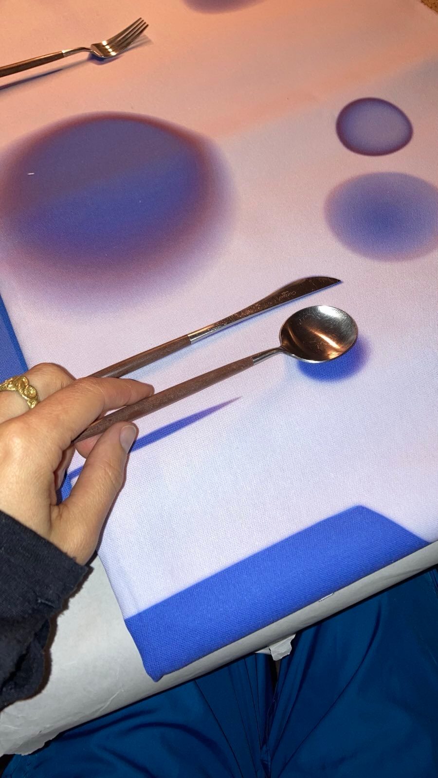 Hand holding spoon and knife, with a fork nearby, on a white tablecloth with blue circular patterns.