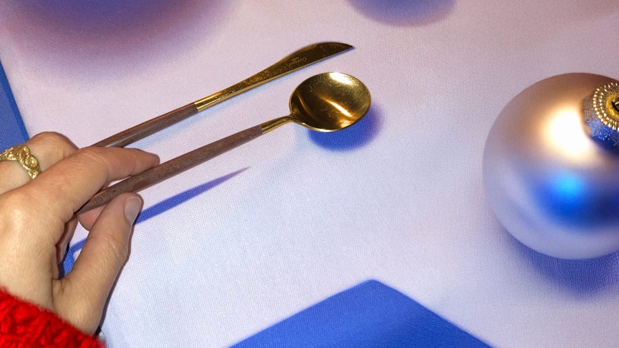 A hand holds golden utensils (spoon, spreader) over blue and white fabric, beside a silver Christmas ornament.