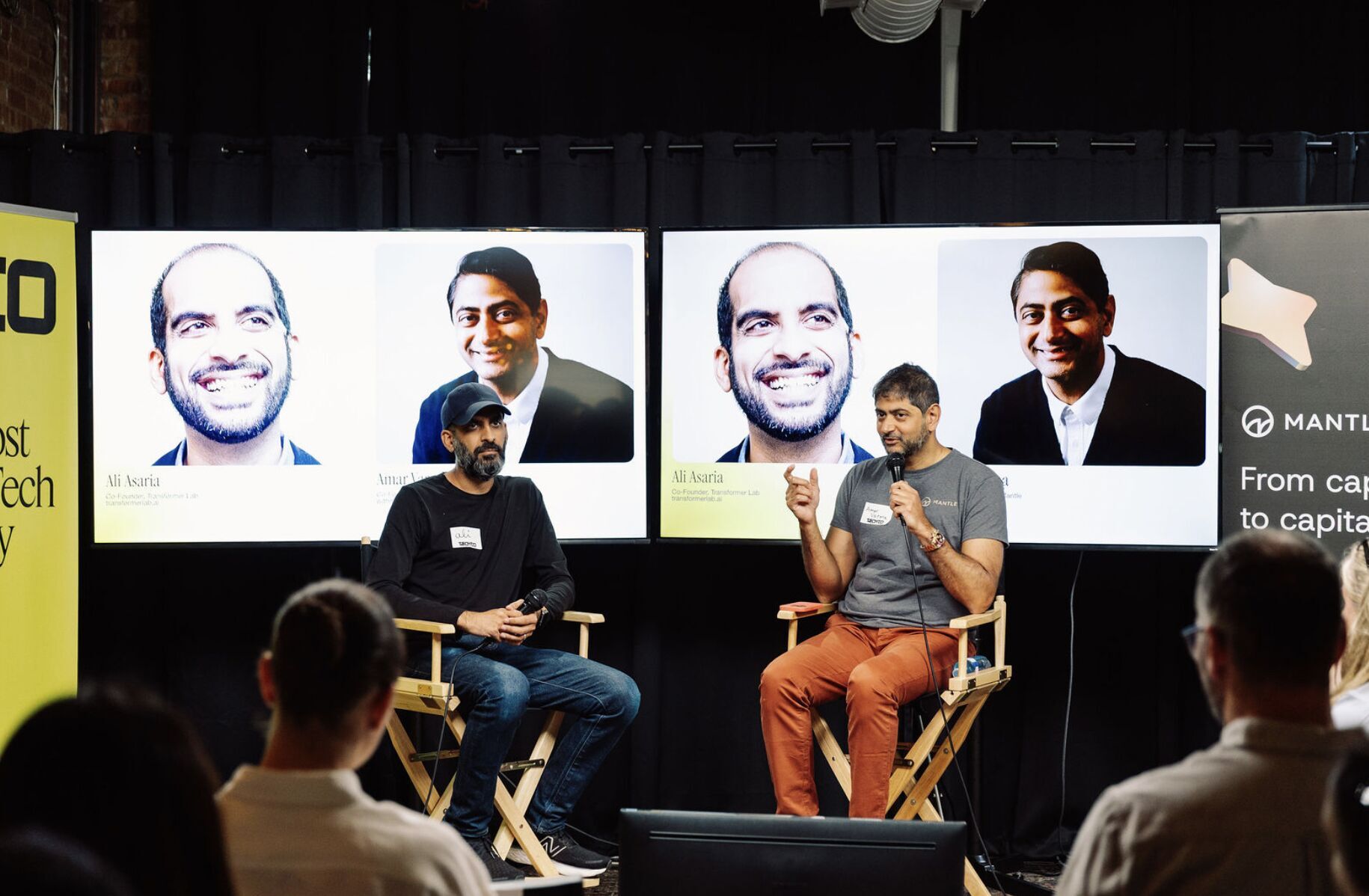 Two men on stage in a panel discussion, one speaking into a microphone, with screens behind.