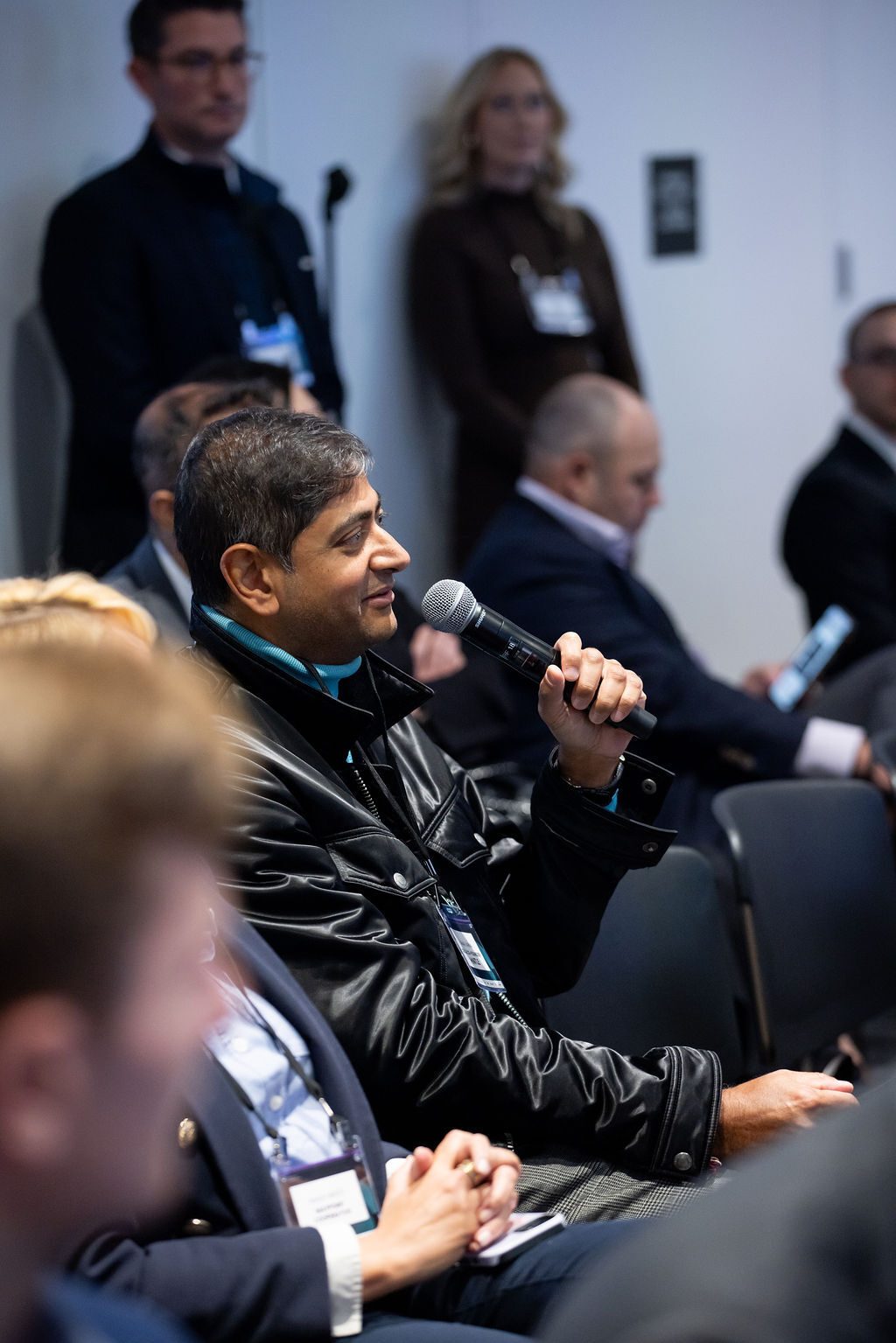Smiling man in black jacket holding microphone, speaking at a conference.