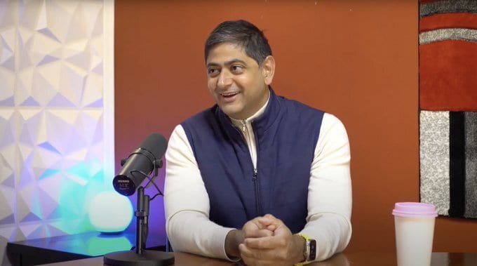 A man in a blue vest smiles while sitting at a desk with a microphone and a cup.