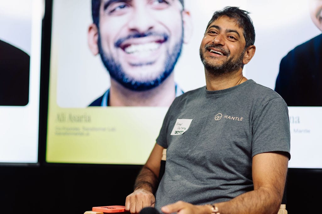 Smiling man in MANTLE t-shirt, name tag, background screen shows another smiling man.