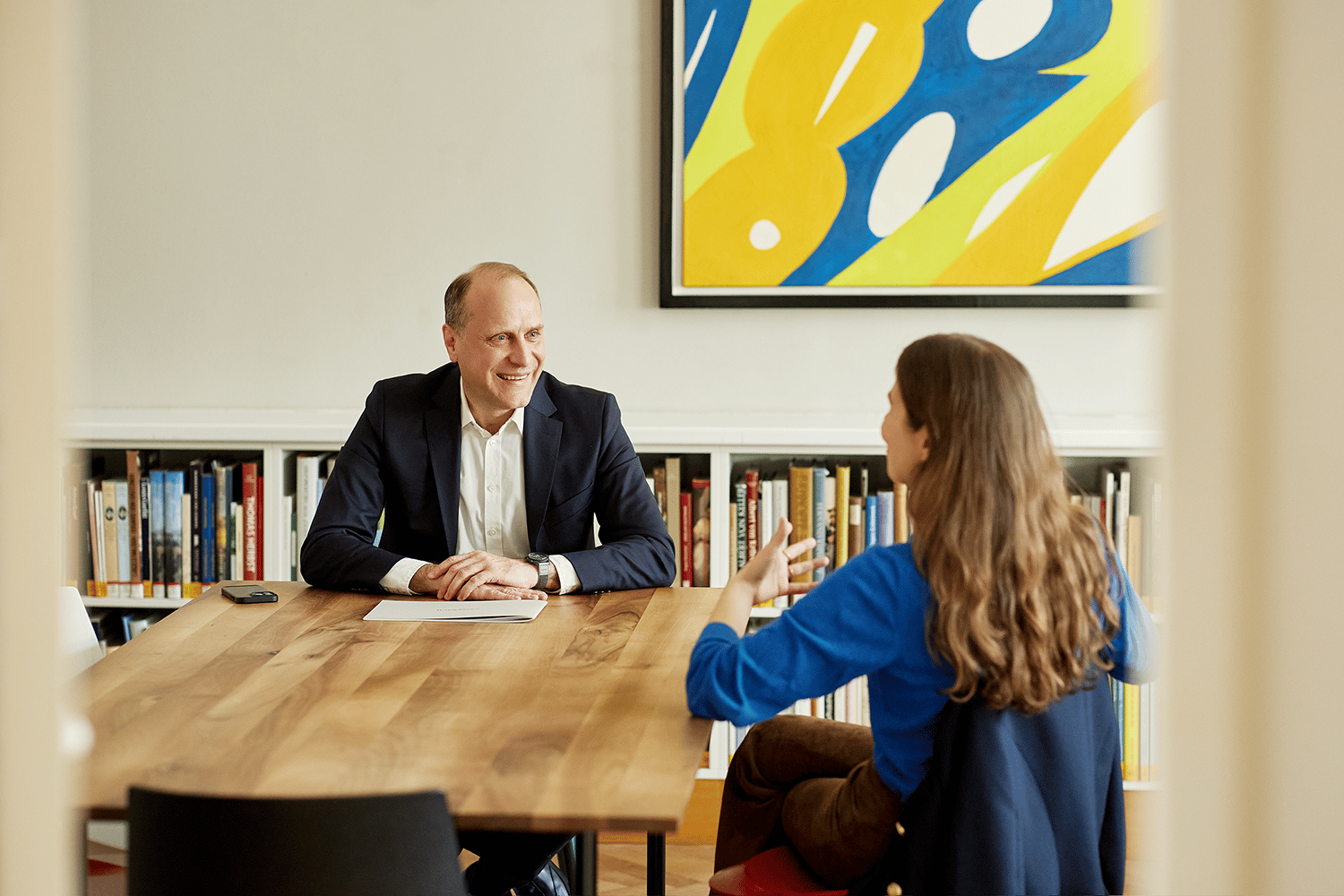 Man in suit smiles at woman gesturing across a table during an office discussion.