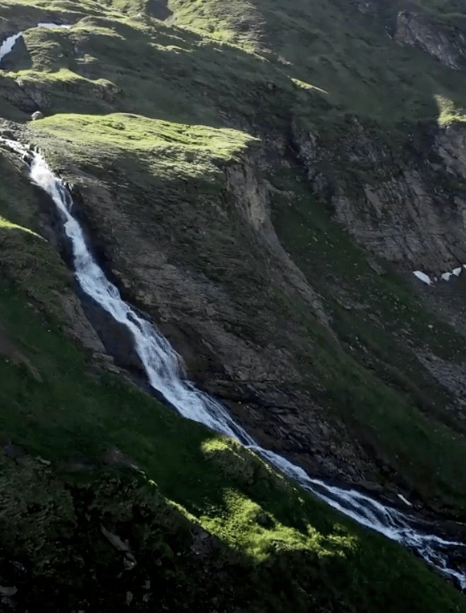 A white waterfall flows down a vibrant green, rocky mountain under sunlight.
