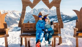 Couple sitting on a decorative wooden bench in snowy mountains under a clear sky, enjoying the view.