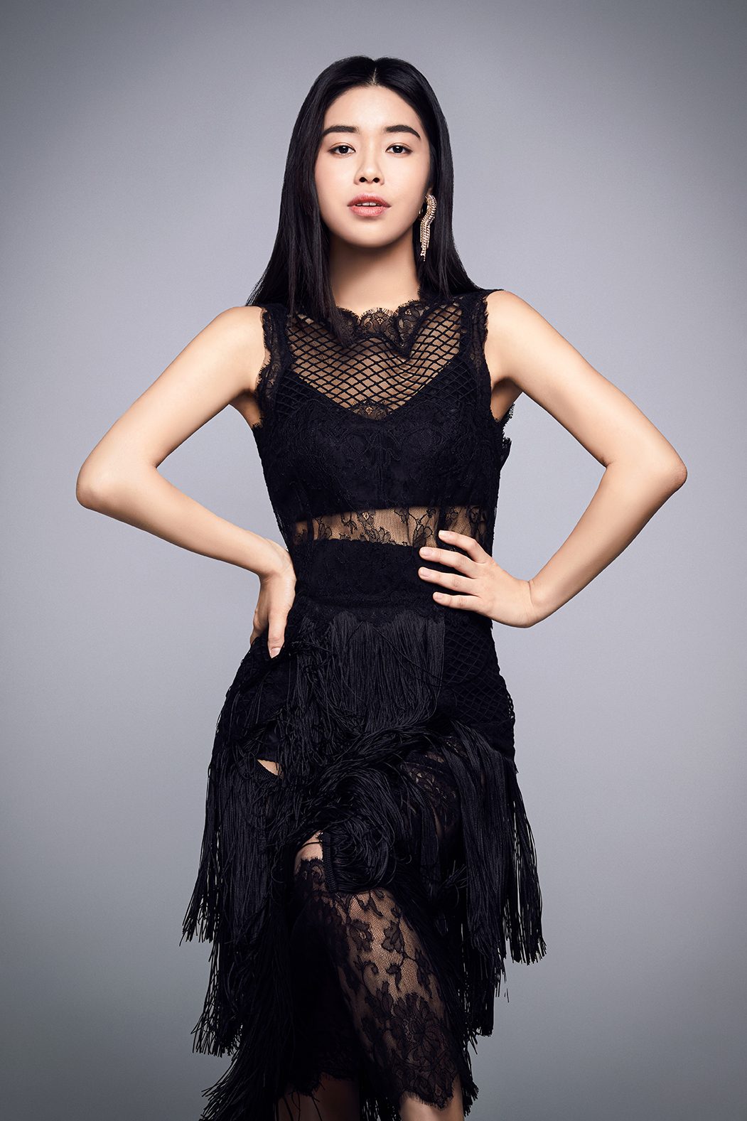 A woman in a black lace and fringe dress, hands on hips, against a grey background.