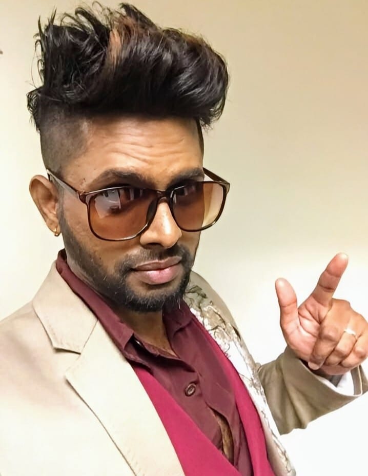 Man with high-volume hair, sunglasses, and beard, wearing a blazer, pointing.