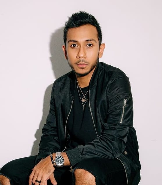 Seated man in black bomber jacket, with necklaces, watch, looking at camera.