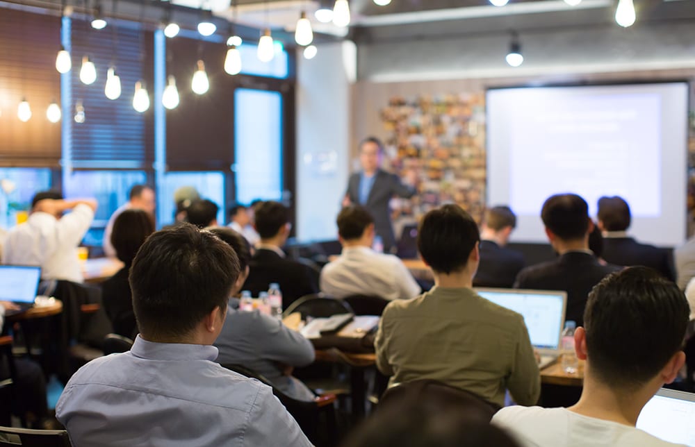 Backs of people in an audience watching a speaker present on a screen.