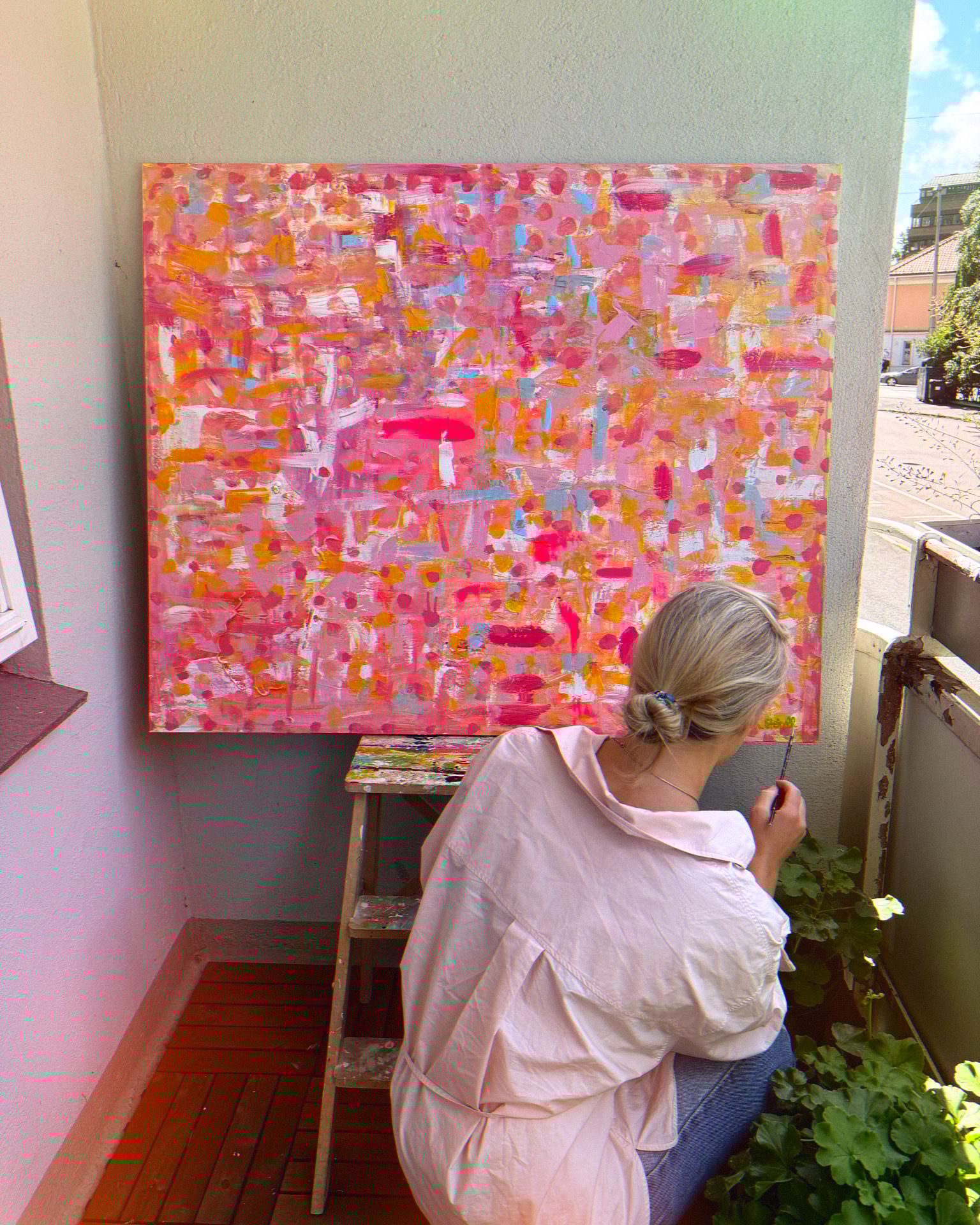 From behind, a person paints a large, colorful abstract canvas on a balcony.