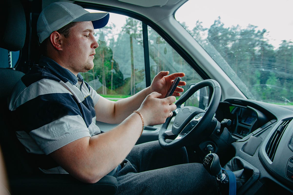 Man in driver's seat, wearing a cap and striped shirt, looking at his phone.