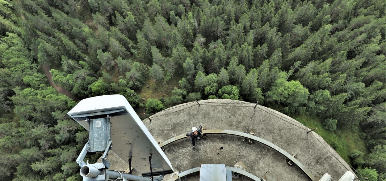 Birds perspective photo of an antenna worker in white helmet peering up at the camera mounted atop the antenna, he is standing on the concrete base of the antenna, amid a sprawling pine forest far below.
