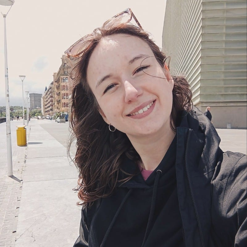 Smiling woman taking a selfie outside with buildings and a street behind her.