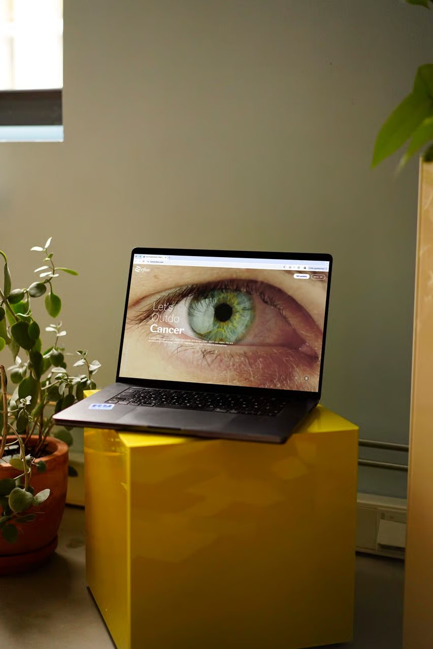 Laptop on yellow cube, screen shows "Let's Outdo Cancer" with eye, Pfizer logo visible.