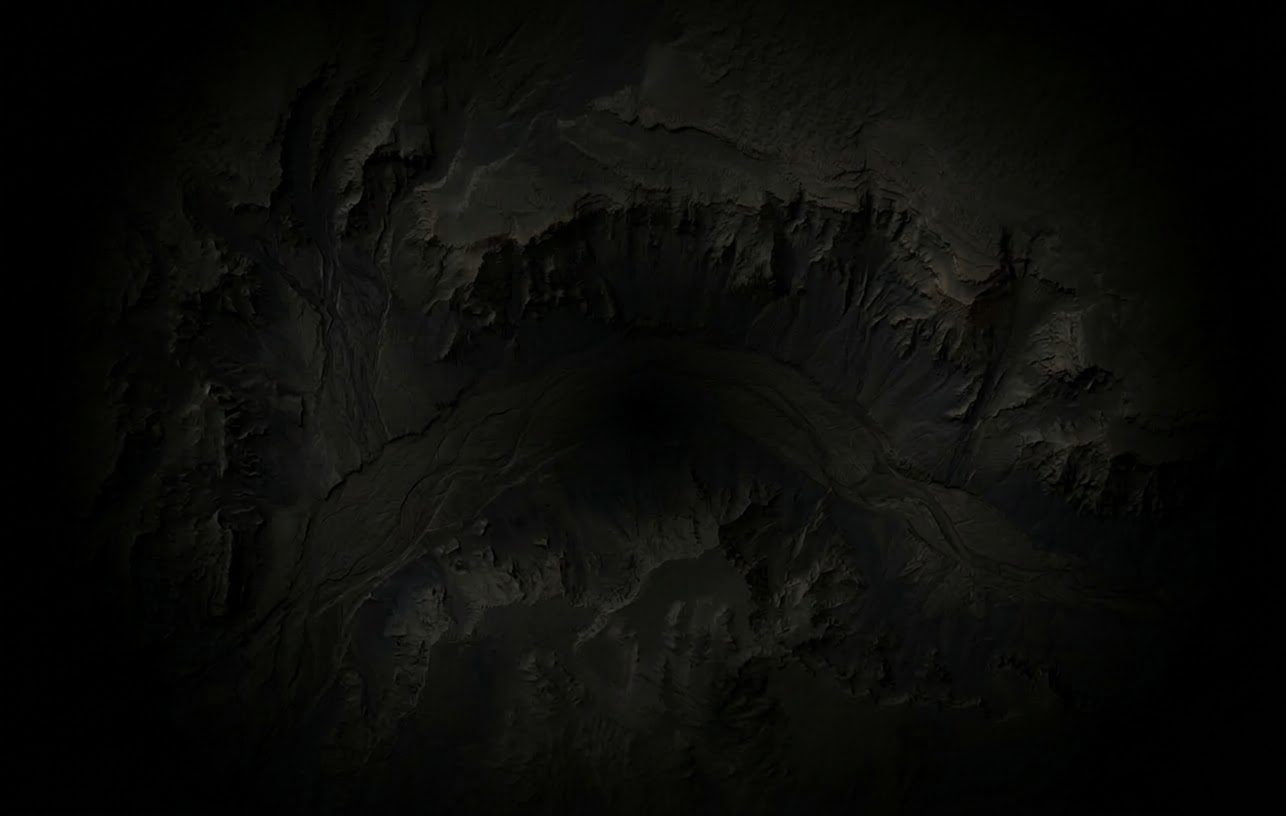 An aerial view of dark, shadowy, rugged terrain with deep canyons and valleys, resembling a monochrome relief map.