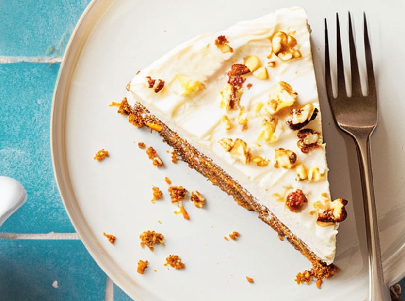 Slice of cheesecake topped with nuts and popcorn, on a white plate with a fork.