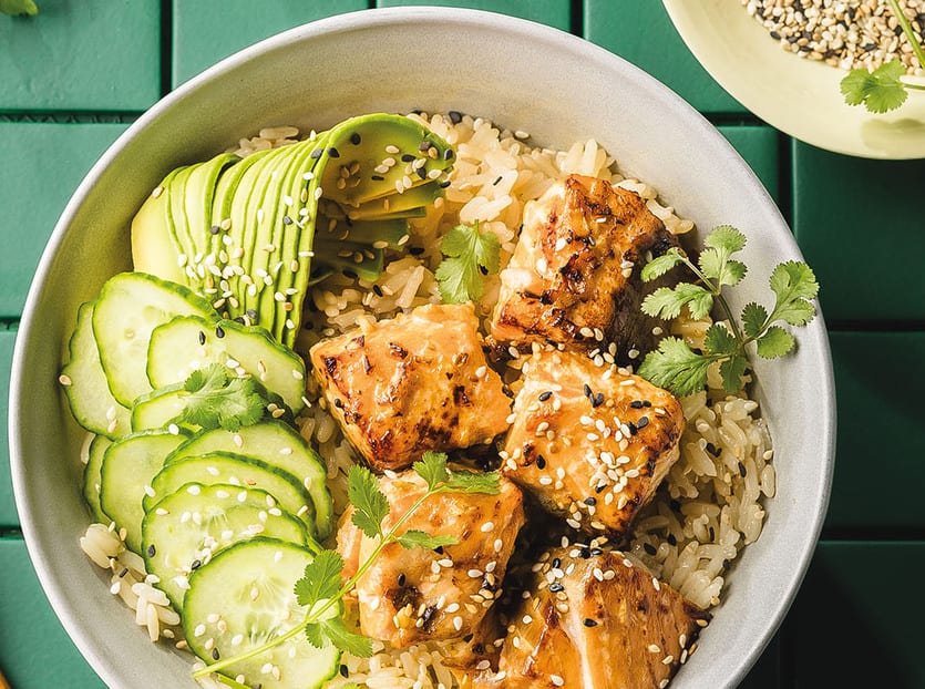 A close-up of a salmon and rice bowl with avocado, cucumber, cilantro, and sesame seeds.