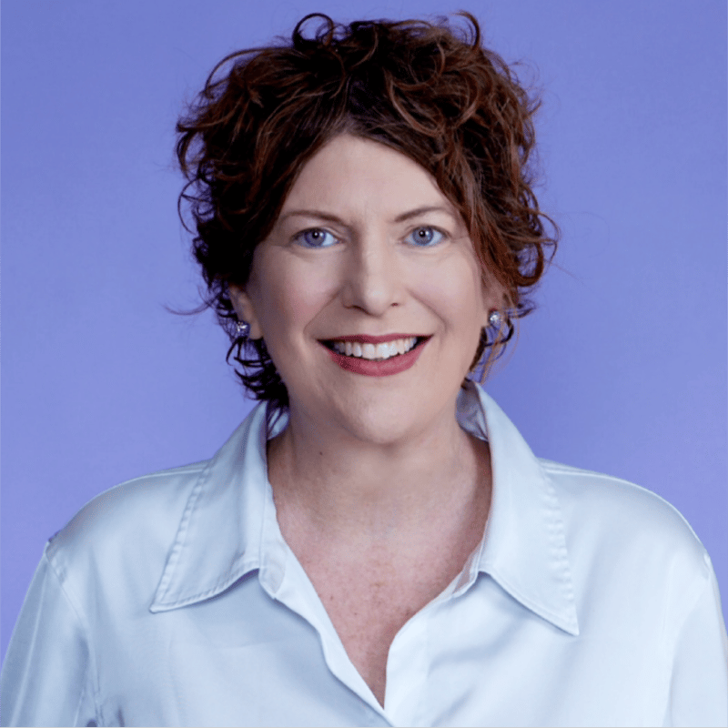 Smiling woman with curly brown hair and blue eyes in a light blue shirt against a purple background.