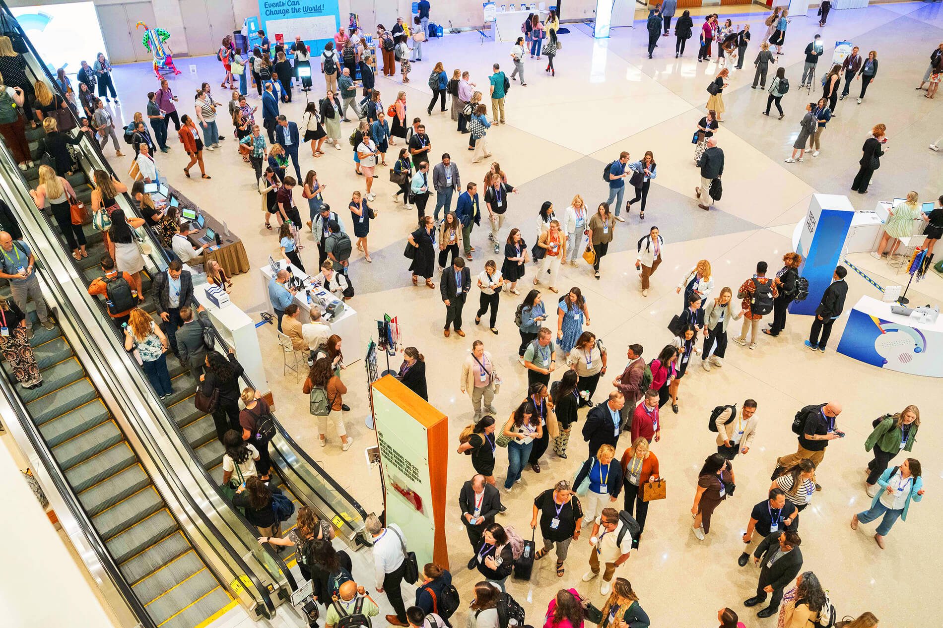 A bustling conference or event hall filled with a large crowd of people walking, congregating, and using escalators.