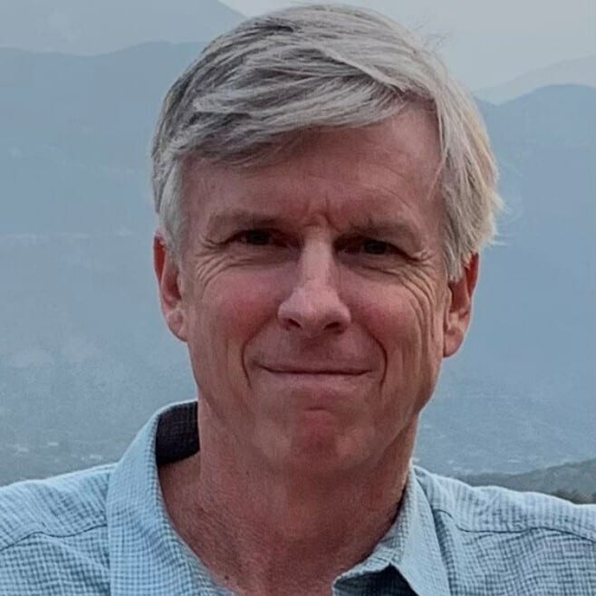A man with gray hair and a light blue checkered shirt smiling, with blurred mountains in the background.