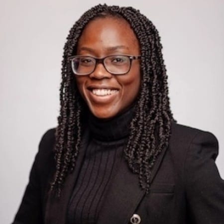 A smiling Black woman with braided hair and glasses, wearing a black turtleneck and blazer.