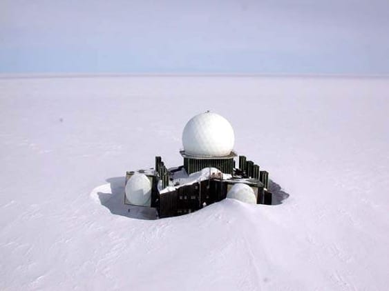 Isolated building with white radomes, partially covered by snow on a vast, flat, white plain.