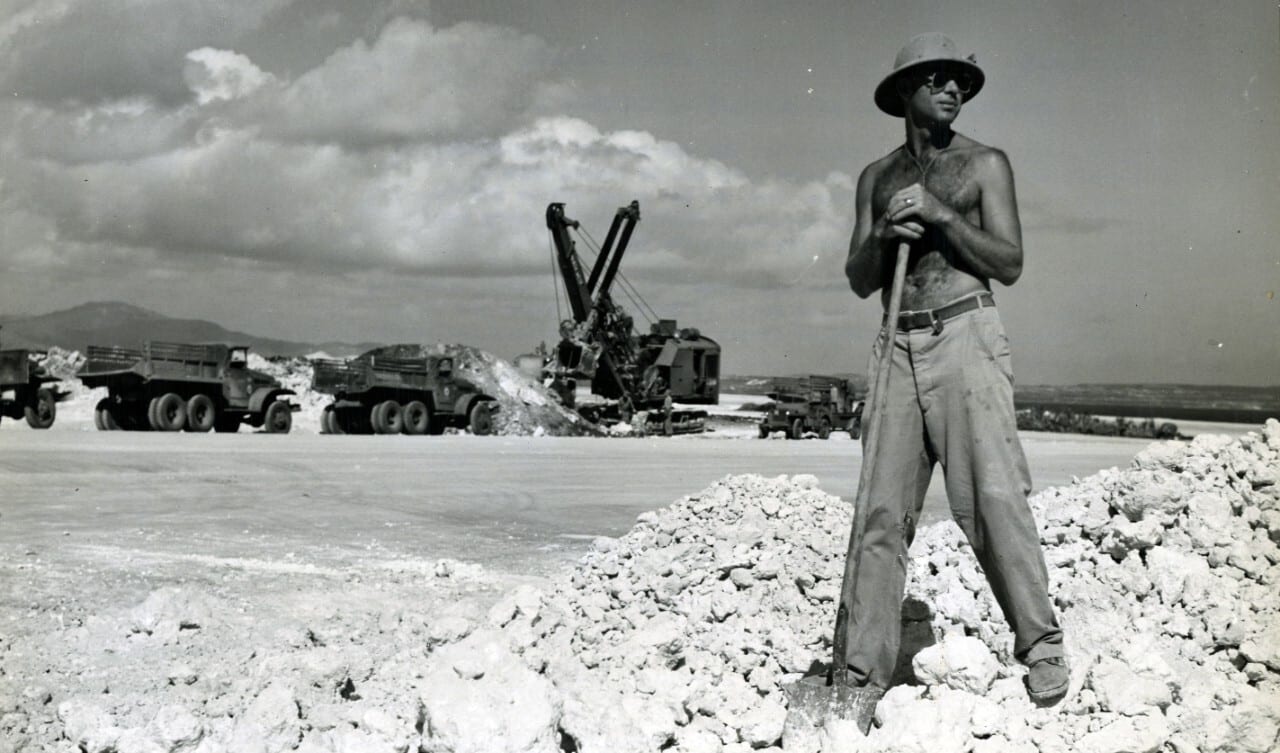Shirtless man with shovel on rock pile at construction site with excavator and trucks in background.