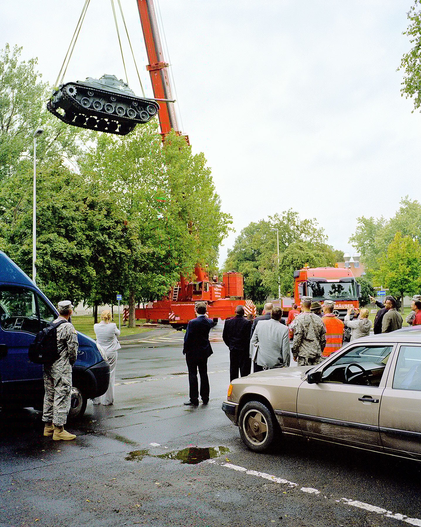 A military tank is lifted by a crane over a street, observed by people and vehicles.