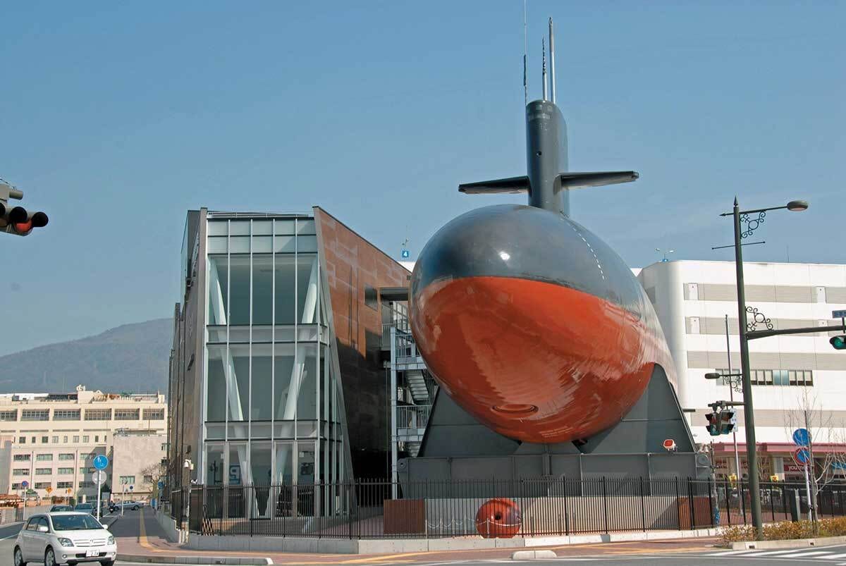Huge grey and red submarine on display beside a modern glass building.