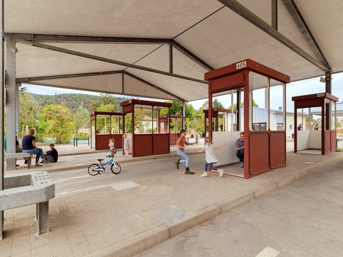 Children play around red bus stop shelters under a large canopy, with adults nearby and a bike.
