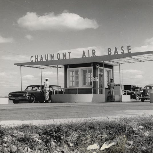 B&W image: Chaumont Air Base entrance gate, guard shack, uniformed men, and cars.